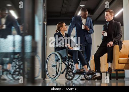 Ritratto a figura intera di un team aziendale diversificato in un ufficio moderno, con una giovane donna in sedia a rotelle che chiacchiera con colleghi maschi. Un ambiente di lavoro inclusivo che mette in evidenza l'accessibilità e il lavoro di squadra. Foto Stock