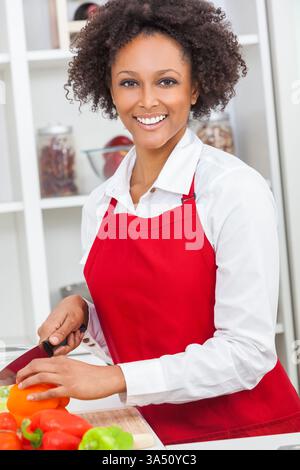 La giovane donna multirazziale che indossa un grembiule rosso trita le verdure in una luminosa cucina casalinga, sorridendo mentre cucina. Un ambiente caldo e di tutti i giorni adatto a ricette, alimentazione sana e pasti in famiglia. Ideale per la cucina domestica e per i contenuti di stile di vita che prevedono la cottura a casa. Foto Stock