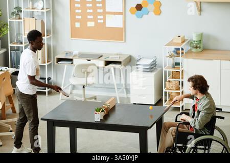 Vista laterale ritratto di giovane con disabilità godendo gioco di ping pong in ufficio sala pausa contro il collega, spazio copia Foto Stock