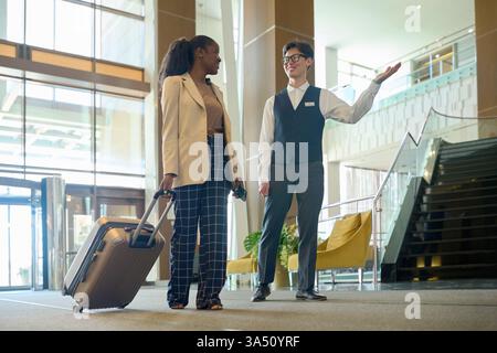 Un receptionist asiatico di sesso maschile che indossa occhiali che mostrano la strada per una donna d'affari di colore che cammina con i bagagli nella lobby dell'hotel Foto Stock