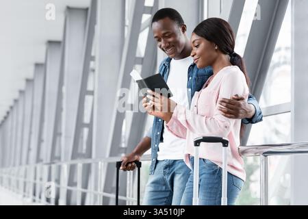 Felice giovane coppia nera in attesa di salire a bordo al terminal dell'aeroporto, controllando insieme i biglietti. Ideale per viaggi, vacanze e romanticismo in scene di viaggio. Questo momento cattura l'attesa, la solidarietà e un'atmosfera di partenza brillante. Foto Stock
