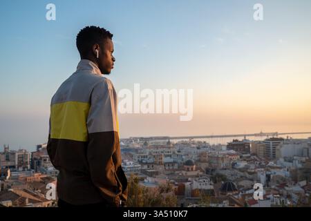 Vista laterale di un giovane uomo sicuro di sé che indossa cuffie senza fili, ammirando una pittoresca città costiera contro un cielo senza nuvole. Perfetto per stili di vita, viaggi e musica in contesti urbani moderni. Atmosfera pulita e rilassata con particolare attenzione alla tecnologia wireless. Foto Stock