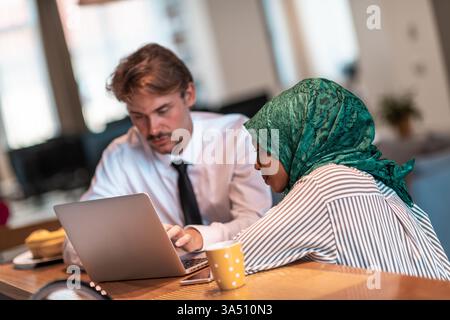 Momenti di lavoro diversificati con una donna musulmana in hijab che collabora con i colleghi utilizzando uno smartphone e un laptop in un moderno ufficio open space. Questa scena multiculturale è ideale per le comunicazioni aziendali, il lavoro di squadra e i contenuti di collaborazione digitale. Foto Stock