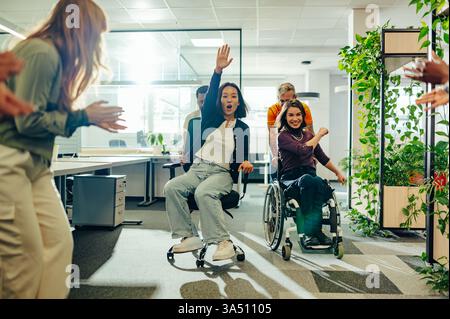 Un team di uffici multirazziali si diverte a gareggiare sulle sedie da ufficio. Una donna asiatica ridendo solleva le braccia mentre passa accanto ai colleghi. Questa immagine mette in evidenza diversità, lavoro di squadra e divertimento sul posto di lavoro. Foto Stock