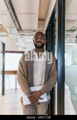 Un uomo afroamericano ben vestito si trova in un moderno ufficio di coworking, con documenti in mano. L'elegante ambiente d'ufficio trasmette professionalità e concentrazione in un ambiente di lavoro contemporaneo. Ideale per i contenuti aziendali, di avvio e d'ufficio, con un professionista sicuro di sé. Foto Stock
