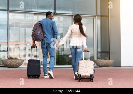 Una coppia nera che si tiene per mano mentre entra in un terminal dell'aeroporto con le valigie. Immagine orientata al viaggio adatta per vacanze, viaggi in famiglia e campagne di viaggio post quarantena o post covid-19. Ideale per le ambientazioni aeroportuali, per le coppie e per i nuovi viaggi. Foto Stock