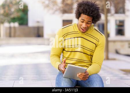 Uomo nero che usa un tablet digitale seduto su una panchina all'aperto. Immagine lifestyle casual e incentrata sulla tecnologia, perfetta per dispositivi mobili, app e contesti di lavoro remoto. Foto Stock
