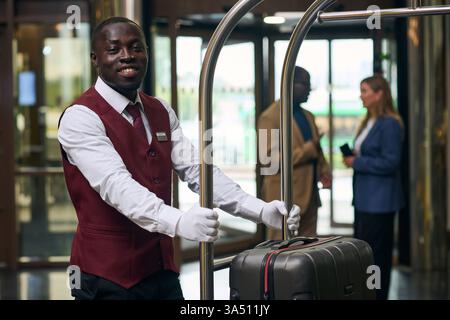 Facchino nero che spinge il carrello con i bagagli mentre cammina vicino agli ospiti Black male e caucasica nella hall dell'hotel Foto Stock