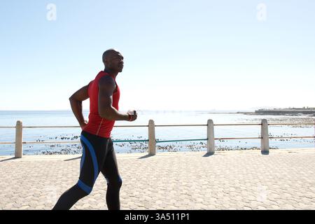 Ritratto di un uomo nero concentrato che fa jogging lungo una passeggiata sul mare in una giornata di sole. Un'immagine versatile per il fitness e lo stile di vita adatta per campagne sportive, attività all'aperto e grafica per vivere in modo sano, con spazio per la copia. Ideale per annunci pubblicitari che promuovono benessere, allenamento e fitness all'aperto diurno. Foto Stock