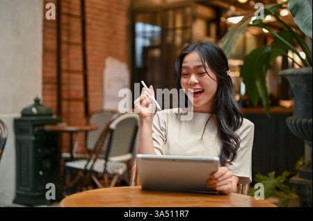 Una bella e felice donna asiatica ha utilizzato il suo tablet digitale e ha avuto una nuova idea per il suo progetto mentre lavorava in remoto in un bar. Vita urbana Foto Stock