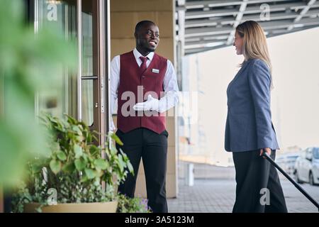 Il fattorino nero dà il benvenuto a una donna caucasica fuori dall'entrata dell'hotel, con bagagli in mano, durante il giorno. Un momento di calda ospitalità che trasmette servizi agli ospiti e check-in senza intoppi. Perfetto per il marchio degli hotel, le scene di ricevimento e le promozioni di viaggio. Foto Stock
