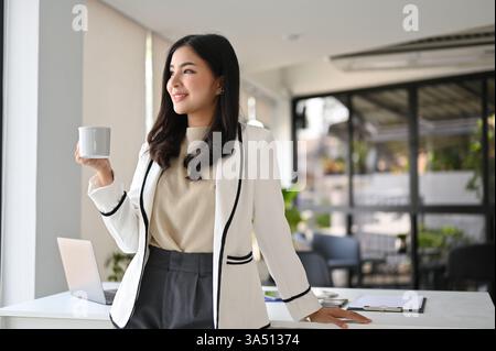 Attraente donna d'affari millenaria asiatica con un vestito bianco si trova nel suo ufficio che guarda fuori dalla finestra. Sogna ad occhi aperti la sua carriera, trasmettendo ambizione e fiducia professionale. Foto Stock