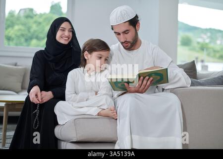 Famiglia musulmana calda e multigenerazionale che legge il Corano durante il Ramadan in una casa moderna. I genitori e il loro figlio si siedono insieme, vestiti con un modesto abbigliamento islamico, condividendo un momento di culto e riflessione. Un ambiente tranquillo e familiare adatto a soddisfare la fede, la tradizione e la solidarietà. Foto Stock