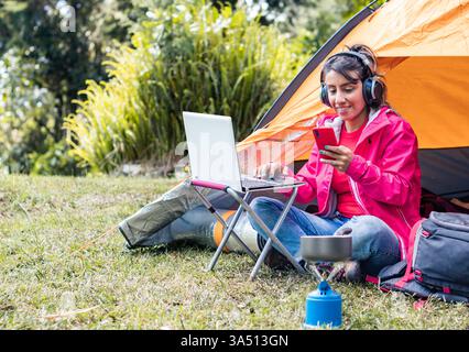 Giovane donna latina all'aperto, multitasking con un laptop e uno smartphone mentre ascolta musica con le cuffie e cucina su una stufa da campeggio. L'immagine cattura in un solo scatto lavori remoti, viaggi e vita all'aperto. Ideale per storie su nomadi digitali, campeggio e automazione dello stile di vita. Foto Stock
