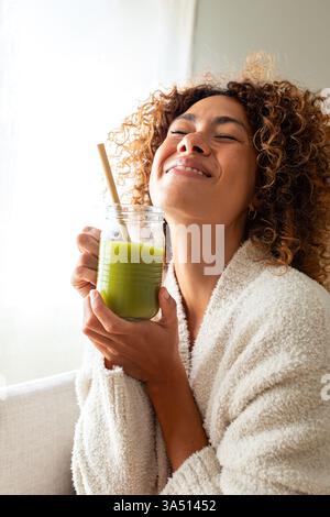Felice donna multirazziale godendo di sano succo verde a casa. Donna che fa disintossicazione bevendo frullato verde. Immagine verticale. Concetto di stile di vita sano. Foto Stock