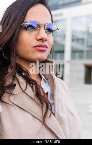 La giovane donna ispanica elegante che indossa un cappotto e un paio di occhiali chic si erge su una strada cittadina con uno sfondo sfocato di grattacieli moderni. Guarda lontano, trasmettendo stile urbano contemporaneo e individualità sicura. Foto Stock
