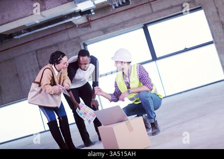 L'architetto discute i piani di progettazione delle case con una giovane coppia multietnica in un cantiere, mostrati su un laptop. Questo ambiente professionale mette in risalto la costruzione di abitazioni, l'architettura e la consulenza ai clienti. Ideale per il settore immobiliare, la progettazione di case e il marketing di costruzioni. Foto Stock