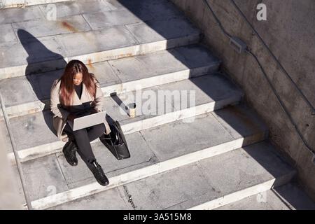 Vista dall'alto di una giovane donna professionista seduta su una scala e che utilizza un notebook, ideale per il lavoro da remoto, l'avvio e i temi aziendali online. Adatto per campagne di tecnologia, imprenditorialità e istruzione che mettono in evidenza la produttività e le competenze digitali. Foto Stock