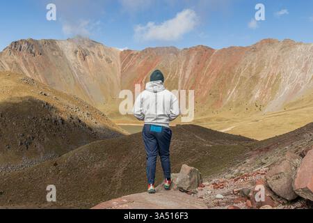 Vista posteriore di un turista maschile irriconoscibile con un berretto e abiti caldi in piedi su una roccia contro il vulcano Nevado de Toluca alla luce del giorno. Suggestivo paesaggio all'aperto, perfetto per viaggi e temi di alpinismo. Ideale per avventure, natura e viaggi in Messico. Foto Stock