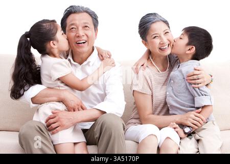 Nipote e nipote cinesi che baciano i loro nonni su un divano in un'accogliente scena familiare multi-generazionale. Un momento emozionante di affetto e di amicizia con la famiglia. Ideale per storie di famiglia, storia e vita familiare. Foto Stock