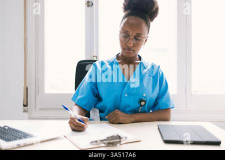 Una dottoressa afroamericana focalizzata in scrub blu scrive appunti su un appunti accanto a un portatile. Questo ritratto professionale trasmette assistenza, lavoro di squadra medico e focus clinico. Foto Stock