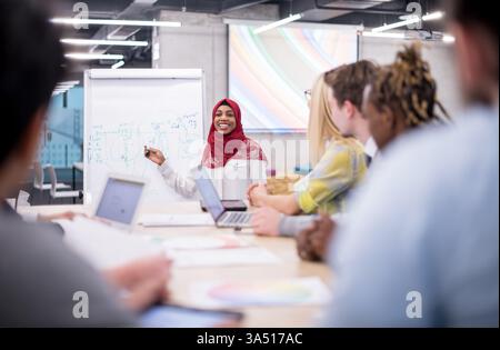 Allegra donna d'affari musulmana con un hijab rosso che conduce una riunione di squadra in un ufficio. Questa immagine di leadership diversificata e sicura funziona bene per le comunicazioni aziendali, i seminari e le campagne di inclusione. Foto Stock
