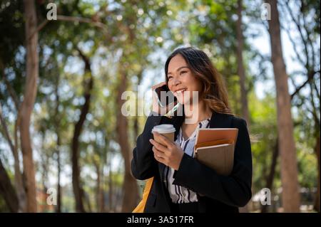 Donna d'affari asiatica positiva che parla al telefono mentre cammina in un parco. Ideale per lavorare in remoto, lavorare e vivere all'aperto in città. Atmosfera luminosa e moderna con tecnologia mobile ed eleganza informale. Foto Stock
