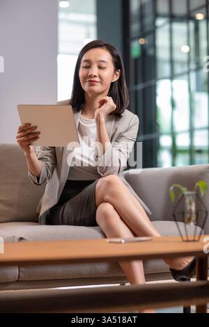 Donna d'affari cinese sicura che usa un tablet in un ufficio moderno. Seduta su un divano, proietta leadership, eleganza e concentrazione. Questa immagine si adatta ad argomenti relativi alle donne nelle aziende, nella tecnologia digitale e nel successo sul posto di lavoro. Foto Stock