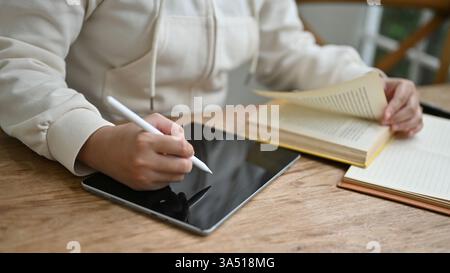 Immagine ravvicinata di una donna con un maglione bianco che usa il suo tablet digitale per gestire le sue attività mentre lavora in remoto in un bar. freelance, college Foto Stock