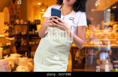 La giovane donna nera sorridente in grembiule e occhiali sfoglia sul suo smartphone appoggiandosi alla porta di un negozio di vetro. L'immagine cattura l'interazione moderna con il retail e lo shopping mobile in un ambiente stradale affollato. Adatto per campagne di vendita al dettaglio, shopping e lifestyle. Foto Stock
