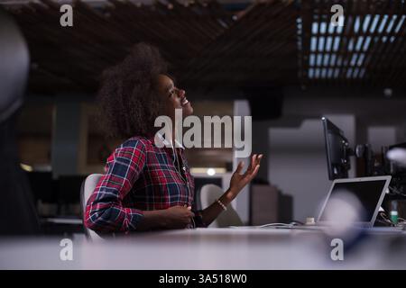 Allegra donna d'affari nera con afro ascolta la musica sugli auricolari e fa un gesto di chitarra a un tavolo. Un ufficio o caffetteria vivace e contemporaneo che mette in risalto la positività e lo stile. Foto Stock