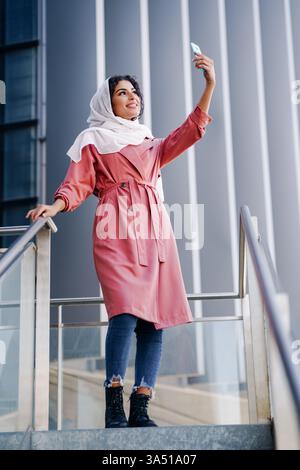 Donna musulmana sorridente in un abito rosa e hijab bianco che si fa un selfie fuori da un ufficio di giorno. Ideale per ambienti di lavoro moderni, social media e immagini di stile di vita con donne musulmane. Foto Stock