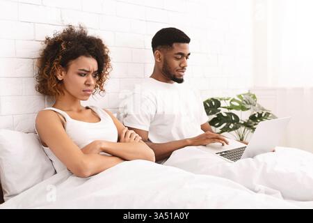Un giovane uomo di colore è sdraiato a letto con un notebook mentre un partner sembra sconvolto, in una stanza da letto che mostra i dispositivi digitali che influenzano le relazioni. Questa scena domestica supporta le discussioni sulla comunicazione online, lo stress digitale e le dinamiche di coppia. Foto Stock