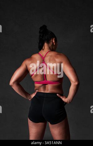 Vista posteriore di un'atleta forte e senza volto con un top rosso e pantaloncini corti sullo sfondo nero. L'immagine enfatizza il fitness, la potenza e un'atmosfera di allenamento stimolante per le campagne di abbigliamento sportivo e benessere. Ideale per il branding in palestra e il marketing del benessere. Foto Stock