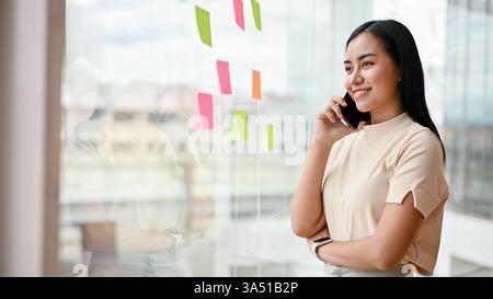 Attraente donna d'affari millenaria asiatica al telefono vicino a una finestra nel suo ufficio. Questo scatto di stile di vita professionale supporta le comunicazioni aziendali, le campagne tecnologiche aziendali e sul luogo di lavoro. Foto Stock