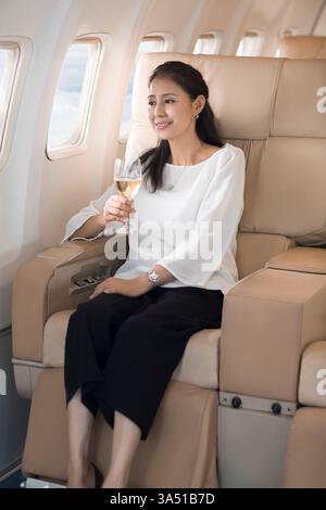 Donna cinese di mezza età sorridente che tiene un bicchiere di vino e guarda fuori dalla finestra mentre si siede sull'aereo Foto Stock