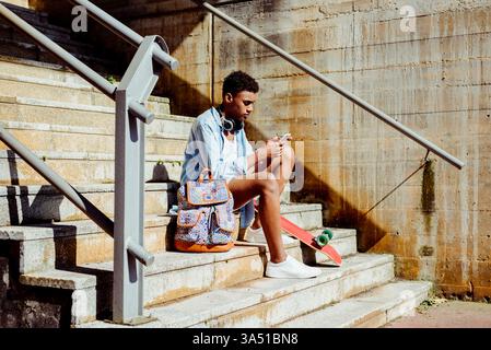Adolescente maschio nero che utilizza uno smartphone sulle scale con uno skateboard e uno zaino nelle vicinanze, catturando lo stile di vita urbano dei giovani. Ideale per campagne tecnologiche, social media e Street style. Foto Stock