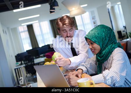 Team aziendale multiculturale che collabora in un ufficio moderno. Una donna musulmana africana in un hijab verde ama il tè mentre una collega mangia una mela accanto ai computer portatili. Questa immagine è adatta alla diversità aziendale, al lavoro di squadra e ai luoghi di lavoro inclusivi. Foto Stock