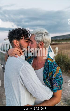 Romantica vista laterale di due giovani uomini che abbracciano e si baciano nella campagna accanto a un furgone retrò. Un caldo momento all'aperto che mette in risalto l'amore, la connessione e l'avventura. Perfetto per lo stile di vita LGBTQ+ e campagne di viaggio con un'atmosfera vintage. Foto Stock