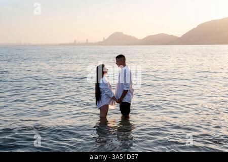 Vista laterale di una coppia latina amorevole che si tiene per mano nel mare al tramonto e si guarda l'un l'altro. Un viaggio romantico e un'immagine di relazione, ideale per vacanze estive, romanticismo e temi di Unione. Foto Stock