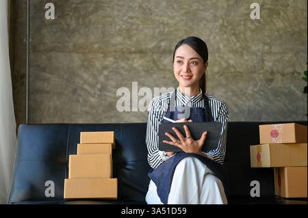 Millennial Asian Woman gestisce un negozio online da un salotto, utilizzando un tablet per gestire gli ordini. Confeziona le scatole per la spedizione in un pratico ufficio domestico, riflettendo il flusso di lavoro di e-commerce delle piccole imprese. Ideale per le imprese femminili, le startup domestiche e le scene di e-commerce in Thailandia. Foto Stock