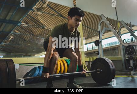 Un uomo asiatico esegue un sollevamento pesante in una palestra vuota. Concentrandosi sulla forza, solleva un barbell con intensità, incarnando potenza e disciplina. Ideale per sollevamento pesi, allenamento in palestra e campagne fitness. Foto Stock