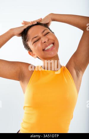 Allegra donna ispanica con i capelli ricci corti che indossano un top giallo e jeans si stagliano su uno sfondo bianco studio. Sorride con gli occhi chiusi e mette le mani sulla testa, proiettando un umore positivo ed espressivo. Foto Stock