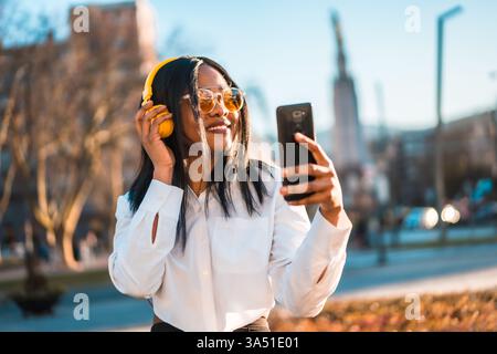 La donna nera sorridente che indossa gli occhiali da sole si siede su una panca del parco al tramonto, ascoltando musica con le cuffie e controllando il suo smartphone. Ottimo per lo stile di vita e per la vita urbana con diverse donne. Cattura momenti rilassati e spensierati e connettività moderna. Foto Stock