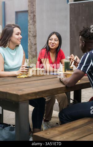 Sorridente donna nativa americana che guarda un'amica caucasica seduta a tavola con gli amici che mangiano insieme in un ristorante all'aperto Foto Stock