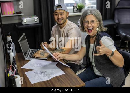Nello studio del tatuaggio, una donna dai capelli grigi siede con un tatuatore che parla di un disegno. L'immagine trasmette la consulenza del cliente e la creazione di body art. Utile per tatuaggi, design, studio e temi del settore della bellezza. Foto Stock