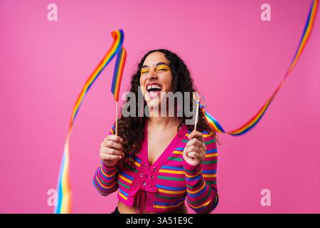 Una allegra donna ispanica con un diastema sorride mentre regge il nastro arcobaleno sullo sfondo di uno studio rosa. La scena colorata mette in risalto l'individualità e l'orgoglio LGBTQ in un ritratto moderno. Ideale per campagne sulla diversità, l'autoaccettazione e la grafica inclusiva. Foto Stock