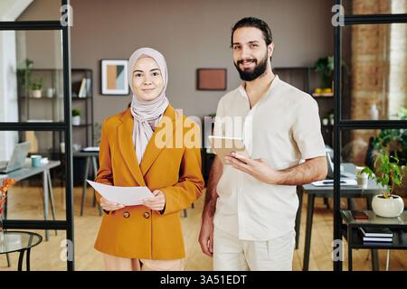Due giovani professionisti, economisti, con un tablet e documenti finanziari in un ufficio open space. La scena diversificata e collaborativa si adatta alle caratteristiche di finanza, analisi e leadership in un ambiente di lavoro moderno. Foto Stock