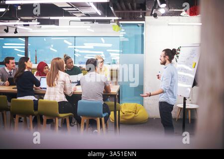 Uomo d'affari multirazziale in piedi e alla guida di una riunione di squadra in un ufficio moderno. Questa immagine mette in evidenza diversità, leadership e collaborazione negli ambienti aziendali. È ideale per il branding aziendale, le comunicazioni di startup e le campagne di lavoro di squadra. Foto Stock