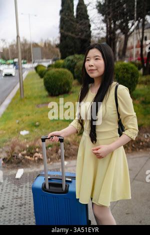 Viaggiatrice asiatica in cerca di un alloggio in città, con bagagli. Questa vivace scena di viaggio mette in evidenza l'esplorazione e la pianificazione, adatta per hotel, guide di viaggio e contenuti sulle destinazioni. Ideale per turismo, promozioni di alloggi e Street style in contesti di viaggio. Foto Stock